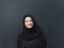 Portrait of beautiful muslim woman in fashionable dress with hijab in front of black chalkboard representing modern islam fashion and ramadan kareem concept photo