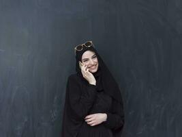 young modern muslim business woman using smartphone wearing sunglasses and hijab clothes in front of black chalkboard photo