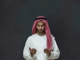 A young Arabian man in traditional clothes making a traditional prayer to God keeps his hands in praying gesture in front of a black background photo