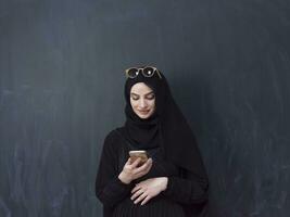 young modern muslim business woman using smartphone wearing sunglasses and hijab clothes in front of black chalkboard photo