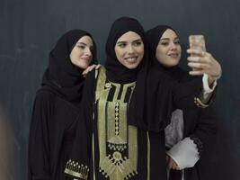 grupo de joven musulmán mujer en de moda vestir con hijab utilizando teléfono inteligente mientras tomando selfie imagen en frente de negro antecedentes foto