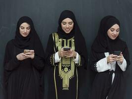 group of beautiful muslim women in fashionable dress with hijab using mobile phone in front of black chalkboard representing modern islam fashion technology and ramadan kareem concept photo