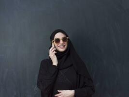 young modern muslim business woman using smartphone wearing sunglasses and hijab clothes in front of black chalkboard photo
