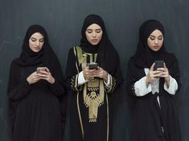 group of beautiful muslim women in fashionable dress with hijab using mobile phone in front of black chalkboard representing modern islam fashion technology and ramadan kareem concept photo