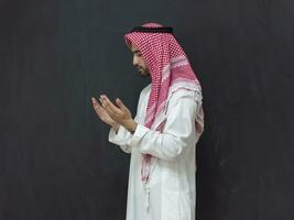 A young Arabian man in traditional clothes making a traditional prayer to God keeps his hands in praying gesture in front of a black background photo