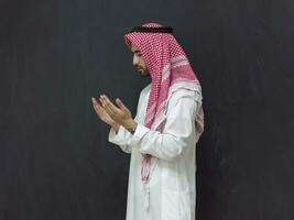 A young Arabian man in traditional clothes making a traditional prayer to God keeps his hands in praying gesture in front of a black background photo