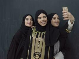 grupo de joven musulmán mujer en de moda vestir con hijab utilizando teléfono inteligente mientras tomando selfie imagen en frente de negro antecedentes foto