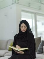 joven tradicional musulmán mujer leyendo Corán en el sofá antes de iftar cena durante un Ramadán banquete a hogar foto