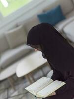 Young traditional muslim woman reading Quran on the sofa before iftar dinner during a ramadan feast at home photo