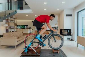 un hombre montando un triatlón bicicleta en un máquina simulación en un moderno vivo habitación. formación durante pandemia condiciones. foto