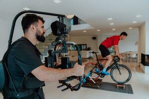 un camarógrafo rodaje un atleta montando un triatlón bicicleta en un simulación máquina en un moderno vivo habitación. formación en pandemia condiciones. foto