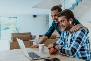 A young married couple is talking to parents, family and friends on a video call via a laptop while sitting in the living room of their modern house photo