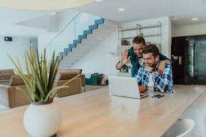 A young married couple is talking to parents, family and friends on a video call via a laptop while sitting in the living room of their modern house photo