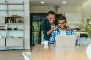 A young married couple is talking to parents, family and friends on a video call via a laptop while sitting in the living room of their modern house photo