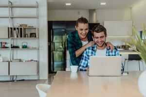 A young married couple is talking to parents, family and friends on a video call via a laptop while sitting in the living room of their modern house photo