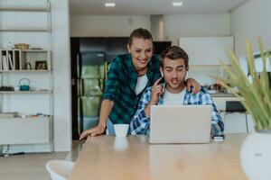 A young married couple is talking to parents, family and friends on a video call via a laptop while sitting in the living room of their modern house photo