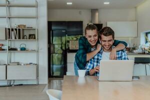 A young married couple is talking to parents, family and friends on a video call via a laptop while sitting in the living room of their modern house photo