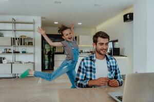 Work from home. Daughter interrupting her father while he is having a business online conversation on his laptop while sitting in modern living room photo