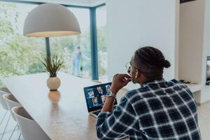 africano americano hombre en lentes sentado a un mesa en un moderno vivo habitación, utilizando un ordenador portátil y teléfono inteligente para negocio vídeo charlar, conversacion con amigos y entretenimiento foto