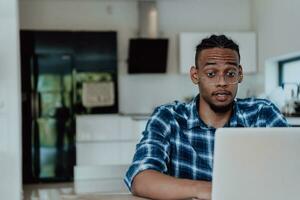 African American man in glasses sitting at a table in a modern living room, using a laptop for business video chat, conversation with friends and entertainment photo