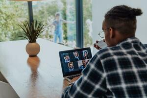 africano americano hombre en lentes sentado a un mesa en un moderno vivo habitación, utilizando un ordenador portátil y teléfono inteligente para negocio vídeo charlar, conversacion con amigos y entretenimiento foto