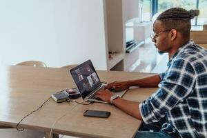 africano americano hombre en lentes sentado a un mesa en un moderno vivo habitación, utilizando un ordenador portátil para negocio vídeo charlar, conversacion con amigos y entretenimiento foto