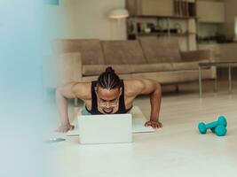 Young African American man working push-ups in the living room while watching online training on laptop photo