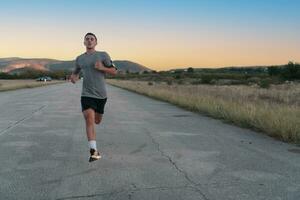 Attractive fit man running fast along countryside road at sunset light, doing jogging workout outdoors photo