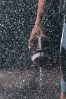 cerca arriba de americano fútbol americano atleta guerrero en pie en un campo atención en pelota y Listo a jugar. jugador preparando a correr, ataque y Puntuación aterrizaje. lluvioso noche con dramático lente llamarada y lluvia gotas. foto