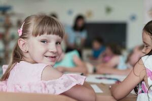 pequeño muchachas sentado en elemental colegio dibujo en papel con su amigos mientras sentado en un moderno salón de clases foto