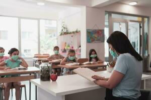 Multiracial group of kids wearing face masks working at class, writing and listening explanations of teacher in classroom photo