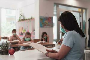 Multiracial group of kids wearing face masks working at class, writing and listening explanations of teacher in classroom photo