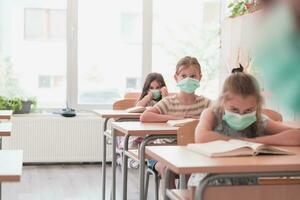 multirracial grupo de niños vistiendo cara mascaras trabajando a clase, escritura y escuchando explicaciones de profesor en salón de clases foto