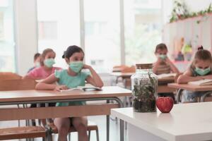 multirracial grupo de niños vistiendo cara mascaras trabajando a clase, escritura y escuchando explicaciones de profesor en salón de clases foto