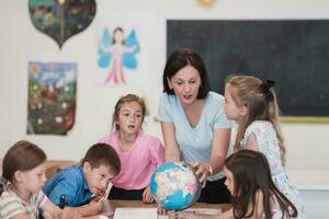 Female teacher with kids in geography class looking at globe. Side view of group of diverse happy school kids with globe in classroom at school. photo