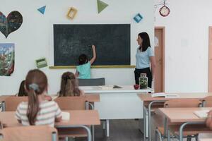 Elementary school. The female teacher helping the child student while writing the answer on the chalkboard. photo