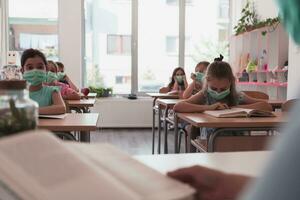 Multiracial group of kids wearing face masks working at class, writing and listening explanations of teacher in classroom photo