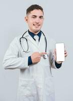 Cheerful doctor holding and pointing at cell phone screen. Young doctor showing and pointing at smatphone screen isolated photo