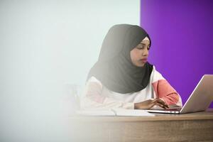 Afro woman wearing a hijab thoughtfully sitting in her home office and uses a laptop and smartphone photo