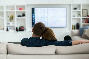 Couple Watches TV together while Sitting on a Couch in the Living Room. Girlfriend and Boyfriend embrace, cuddle, talk, smile and watch Television Streaming Services. Home with Cozy Stylish Interior. photo