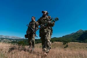 A sniper team squad of soldiers is going undercover. Sniper assistant and team leader walking and aiming in nature with yellow grass and blue sky. Tactical camouflage uniform. photo