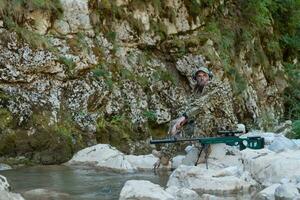 A military man or airsoft player in a camouflage suit sneaking the river and aims from a sniper rifle to the side or to target. photo