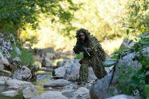 A military man or airsoft player in a camouflage suit sneaking the river and aims from a sniper rifle to the side or to target. photo