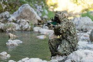 un militar hombre o airsoft jugador en un camuflaje traje furtivo el río y objetivos desde un francotirador rifle a el lado o a objetivo. foto