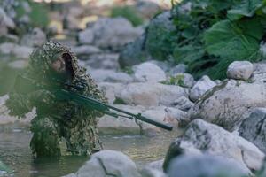 A military man or airsoft player in a camouflage suit sneaking the river and aims from a sniper rifle to the side or to target. photo