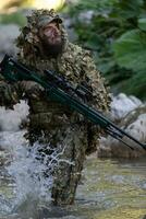 A military man or airsoft player in a camouflage suit sneaking the river and aims from a sniper rifle to the side or to target. photo