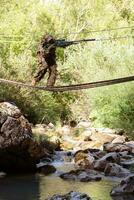 A military man or airsoft player in a camouflage suit sneaking the rope bridge and aims from a sniper rifle to the side or to target. photo