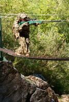 A military man or airsoft player in a camouflage suit sneaking the rope bridge and aims from a sniper rifle to the side or to target. photo