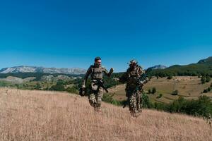 A sniper team squad of soldiers is going undercover. Sniper assistant and team leader walking and aiming in nature with yellow grass and blue sky. Tactical camouflage uniform. photo