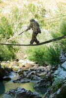 A military man or airsoft player in a camouflage suit sneaking the rope bridge and aims from a sniper rifle to the side or to target. photo